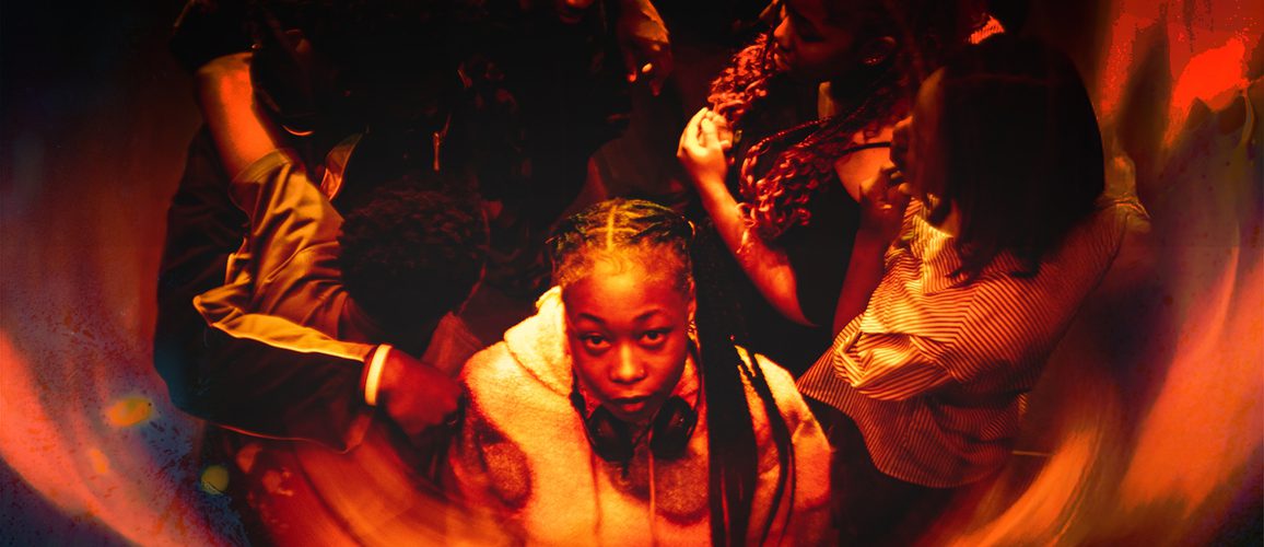 A photo in a red and orange hue showing a group of teenagers stood in a circle with one stood at the front staring at the camera. It feels as if they are being engulfed by flames.