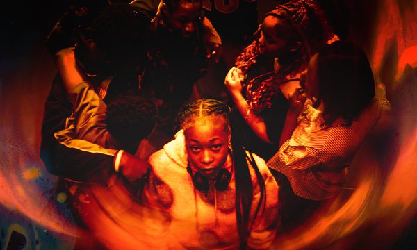 A photo in a red and orange hue showing a group of teenagers stood in a circle with one stood at the front staring at the camera. It feels as if they are being engulfed by flames.