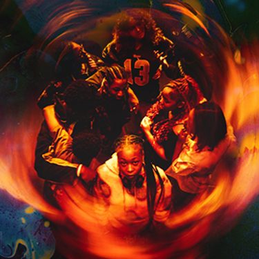 A photo in a red and orange hue showing a group of teenagers stood in a circle with one stood at the front staring at the camera. It feels as if they are being engulfed by flames.