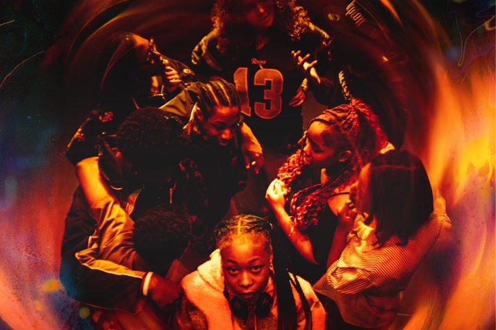 A photo in a red and orange hue showing a group of teenagers stood in a circle with one stood at the front staring at the camera. It feels as if they are being engulfed by flames.