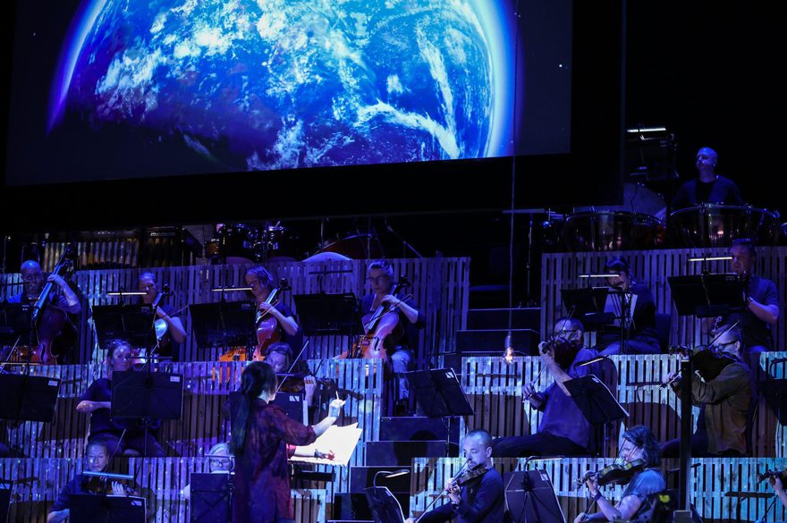 The back of a conductor wearing red is seen with an orchestra playing behind in blue light and a blue moon above.