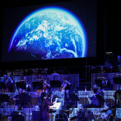 The back of a conductor wearing red is seen with an orchestra playing behind in blue light and a blue moon above.
