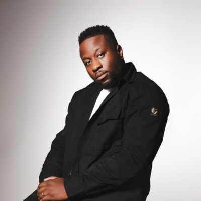 Babatunde Aléshé is sitting against a grey wall weairng a black suit. He's looking straight into the camera with his hand crossed over in his lap.