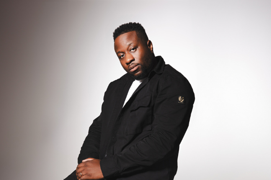 Babatunde Aléshé is sitting against a grey wall weairng a black suit. He's looking straight into the camera with his hand crossed over in his lap.