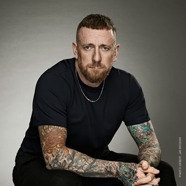 Bradley Wiggins is wearing black short sleevd top and black trousers and is sitting down slouching forward with his hands clasped together in front of him.