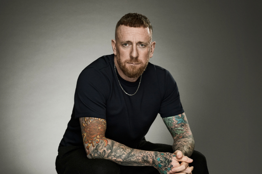 Bradley Wiggins is wearing black short sleevd top and black trousers and is sitting down slouching forward with his hands clasped together in front of him.