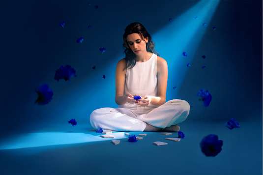 A woman in white sits cross-legged on the floor under a blue spotlight, holding a blue flower, with flower petals and white fragments scattered around her.