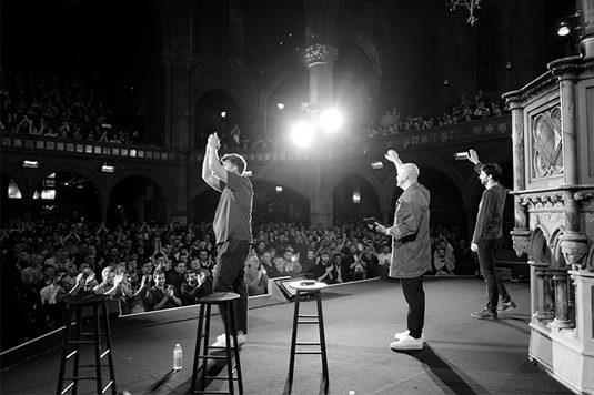 A photo taken from behind the three hosts of Football Cliches Live as they stand on a stage looking out to the audience clapping.