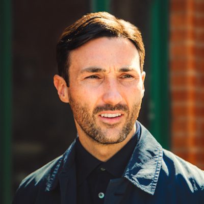Josh is tall, dark hair standing in the sunshine next to orange brick wall with dark air and navy blue shirt and jacket.
