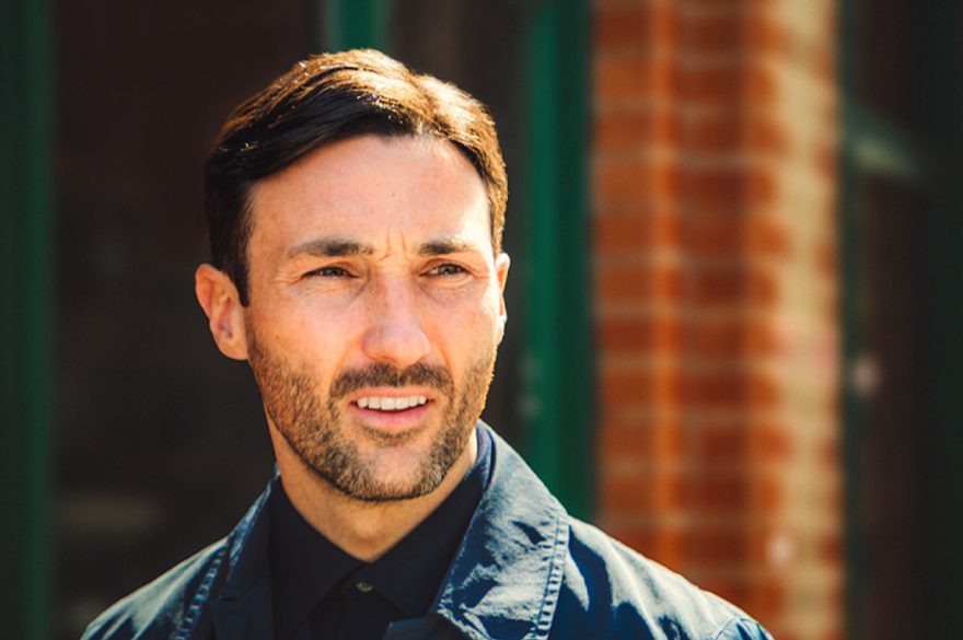Josh is tall, dark hair standing in the sunshine next to orange brick wall with dark air and navy blue shirt and jacket.