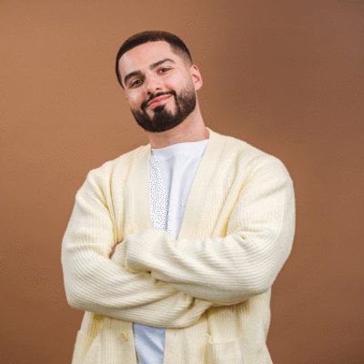 Kae Kurd has short brown hair and a beard and is smiling with his arms crossed against his chest. He is wearing a white shirt and a cream-colored cardigan against a plain brown background.