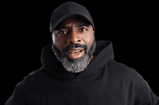 Kane Brown is wearing a black hoodie and cap and is looking straight into the camera with a surprise smirk on his face