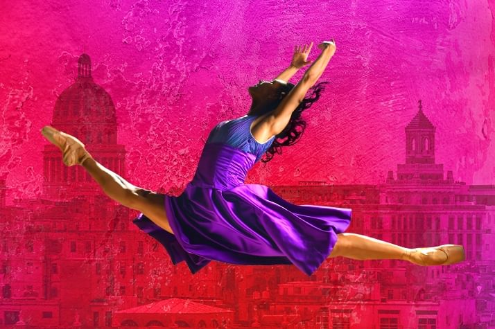 A ballet dancer wearing a purple gown is in mid air in a jump split with their arms above their heads. Behind them is a city background in red/pink.