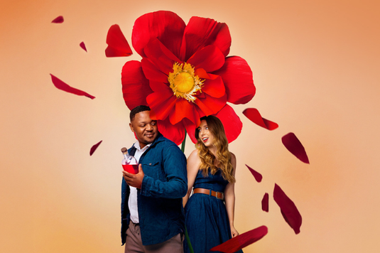 A man holding a drink and a woman stand back to back in front of a large red flower with petals floating around them against a beige background.