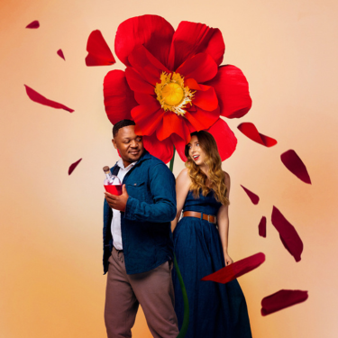 A man holding a drink and a woman stand back to back in front of a large red flower with petals floating around them against a beige background.