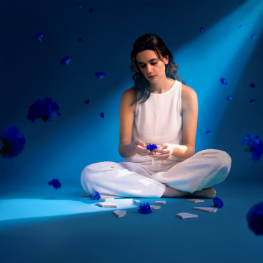 A woman in white sits cross-legged on the floor under a blue spotlight, holding a blue flower, with flower petals and white fragments scattered around her.