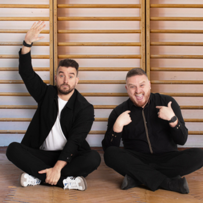 The brothers are in all black and are sitting down on the classroom floor with their legs crossed, while smiling towards the camera.