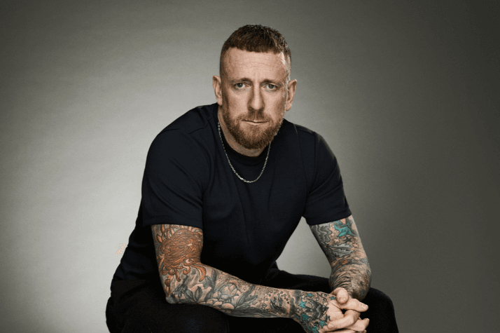 Bradley Wiggins is wearing black short sleevd top and black trousers and is sitting down slouching forward with his hands clasped together in front of him.
