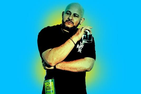 White Yardie is wearing a black top and is standing up straight with his arms crossed against his chest while holding a glass in one hand.