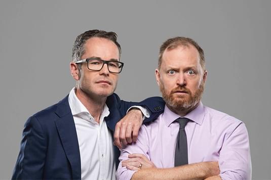 Two men in suits standing side by side. one is in a navy suit with his hand on there other males shoulder with a straight look on his face, while the other guy is in a pink shirt and has a shocked expression on his face.