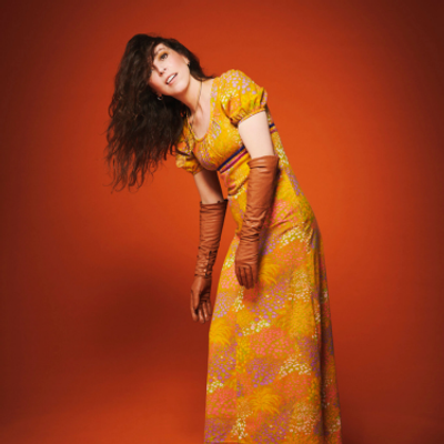 Bridget Christie is wearing a yellow dress and glove and is stood leaning with some of her hair over her head against a red background
