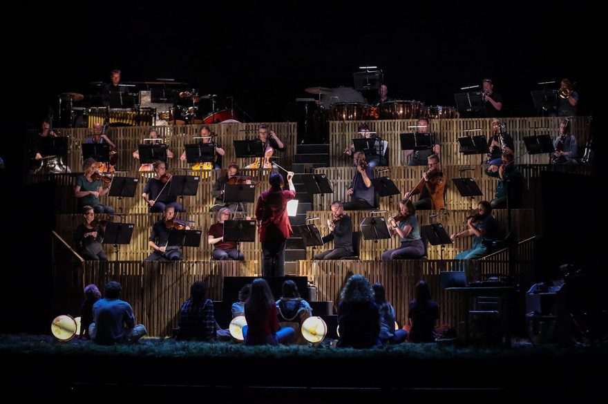 An orchestra is playing on raised rostras with the back of children sitting and watching them from the stage.