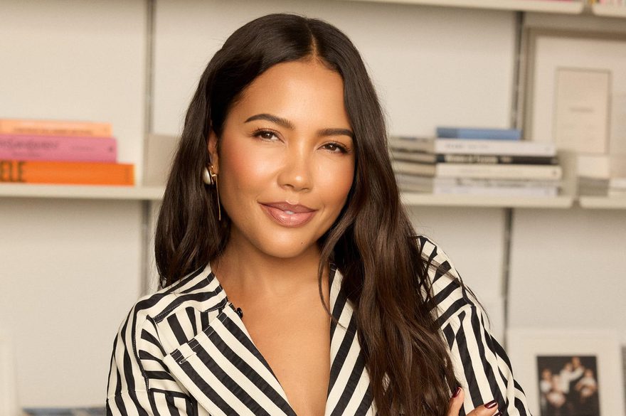 Emma Grede leans on a desk with her arms crossed in a striped shirt and black spotted trousers.