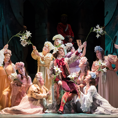 An ensemble groups together on stage, all holding white flowers in the air. They are dressed in period clothing and some stand, while others sit.