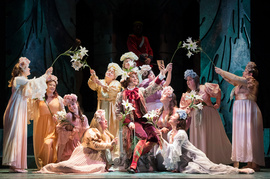 An ensemble groups together on stage, all holding white flowers in the air. They are dressed in period clothing and some stand, while others sit.