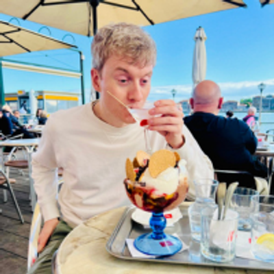 James Acaster is sitting at a table, looking at a dessert placed in front of him.