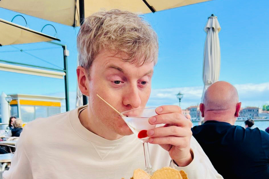 James Acaster is sitting at a table, looking at a dessert placed in front of him.