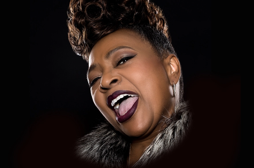 A close-up studio portrait of Jocelyn Brown mid-performance. She is singing with her mouth open wide, showing strong emotion and energy. Her head is tilted slightly back, eyes partly closed, conveying power and joy. She has short, styled hair with volume on top, wears dark lipstick, subtle eye makeup, hoop earrings, and a fur-textured collar or jacket. The background is black, drawing focus to her expressive face and voice in the moment.