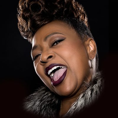 A close-up studio portrait of Jocelyn Brown mid-performance. She is singing with her mouth open wide, showing strong emotion and energy. Her head is tilted slightly back, eyes partly closed, conveying power and joy. She has short, styled hair with volume on top, wears dark lipstick, subtle eye makeup, hoop earrings, and a fur-textured collar or jacket. The background is black, drawing focus to her expressive face and voice in the moment.