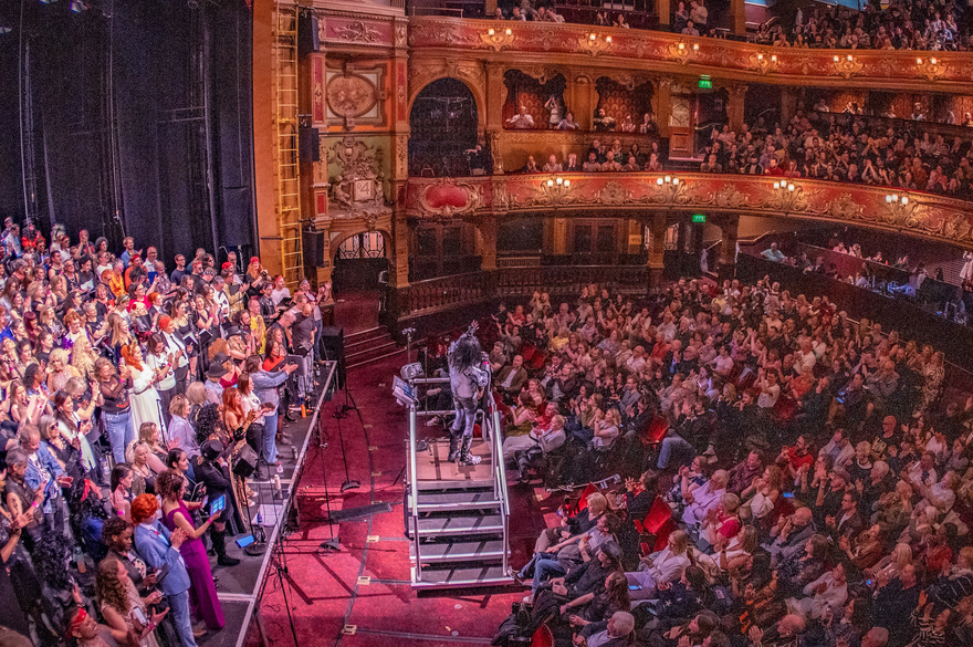 A packed theatre audience, with many people engaged in a live performance on stage at Hackney Empire