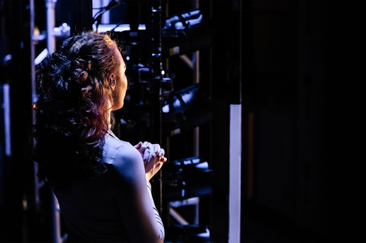 A woman with curly red hair stands in the wings of a theatre, with her hands held together against her chest. Lighting equipment can be seen next to her.
