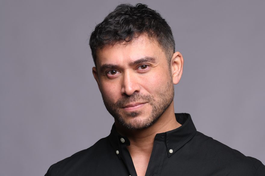 Rav wilding is wearing a black short with a red Ralph Lauren logo on the chest area and is looking straight into the camera whle standing agaisnt a light grey wall.