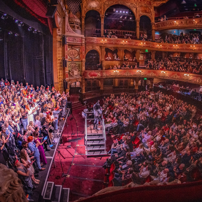 A packed theatre audience, with many people engaged in a live performance on stage at Hackney Empire