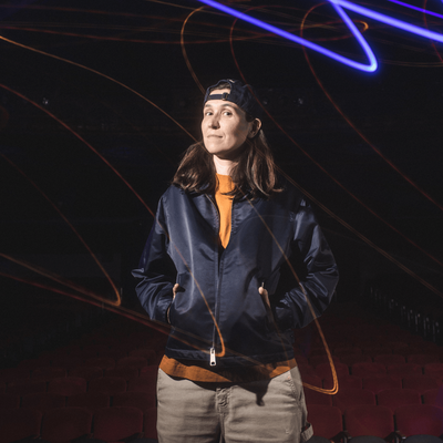 Ashley Gavin stands in a jacket and hat poses in front of a glowing light, highlighting her features and creating a soft ambiance.