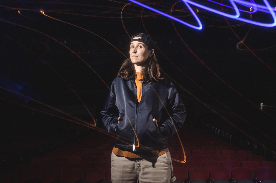 Ashley Gavin stands in a jacket and hat poses in front of a glowing light, highlighting her features and creating a soft ambiance.
