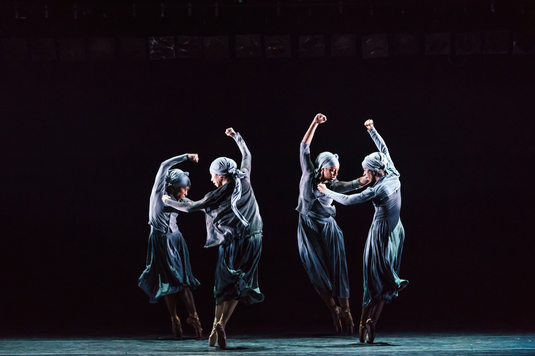 A group of four dancers wearing blue dresses elegantly performing together on stage in a vibrant setting.