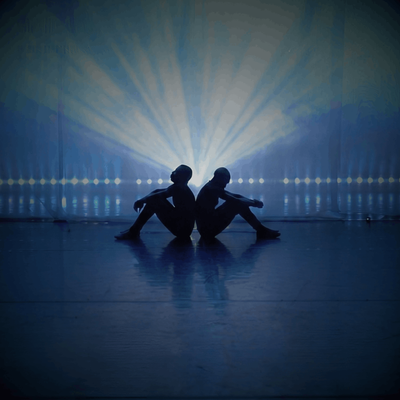 The image shows two people sitting on the floor, back-to-back, in the centre of stage. Their bodies are in silhouette, so you can’t see their facial features or clothing details—only their outlines. Both have their knees bent and arms resting loosely, giving a calm, reflective posture. Behind them, a very bright light shines directly toward the camera, creating dramatic rays that spread outward like a fan. This strong backlight makes the figures appear almost like shadows against a glowing blue background.
