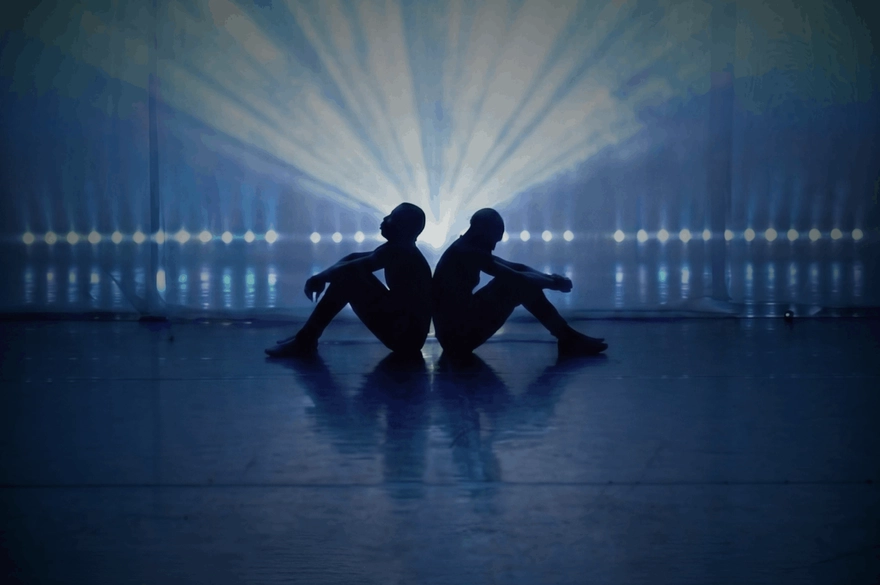 The image shows two people sitting on the floor, back-to-back, in the centre of stage. Their bodies are in silhouette, so you can’t see their facial features or clothing details—only their outlines. Both have their knees bent and arms resting loosely, giving a calm, reflective posture. Behind them, a very bright light shines directly toward the camera, creating dramatic rays that spread outward like a fan. This strong backlight makes the figures appear almost like shadows against a glowing blue background.