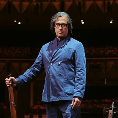David Olusoga is wearing a blue buttoned shirt and is standing up straight holding a wooden stick in his hand looking straight into the camera.