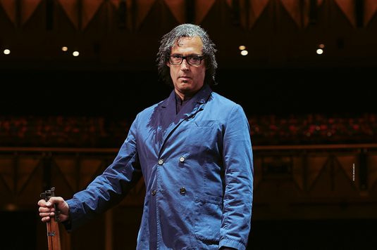 David Olusoga is wearing a blue buttoned shirt and is standing up straight holding a wooden stick in his hand looking straight into the camera.