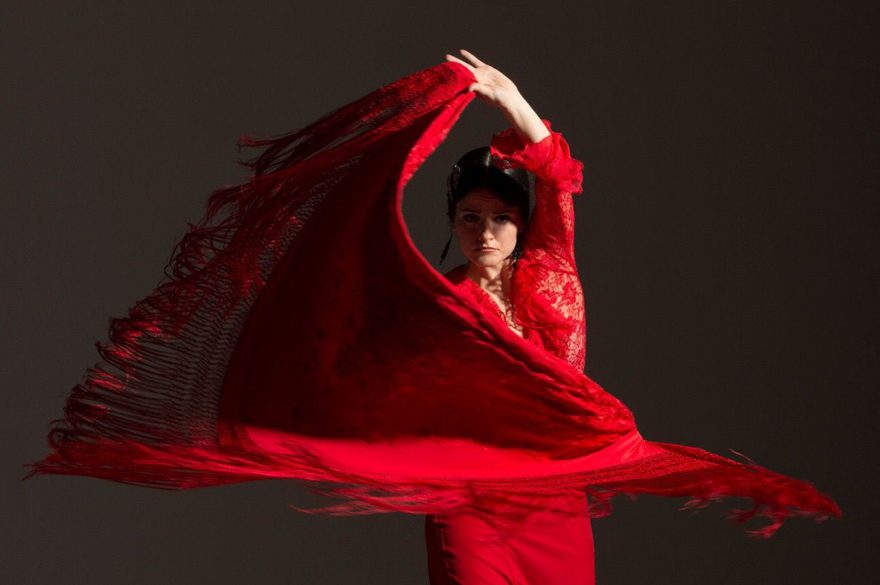 A female Flamenco dancer with dark hair, wearing a long, bright red dress with lace top and ruffles at the bottom of her skirt is standing and swirling a large red shawl with long fringes around her. Her left arm is raised above her head, holding one end of the shawl. She is lit by an overhead spotlight from her left on a dark grey background.