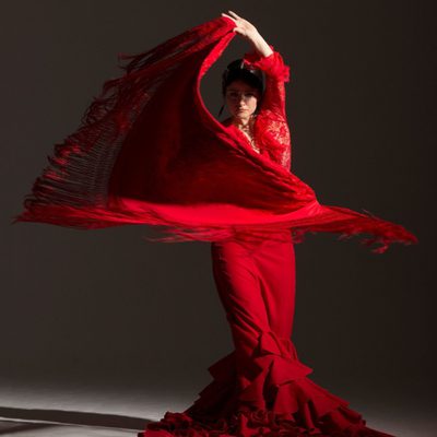 A female Flamenco dancer with dark hair, wearing a long, bright red dress with lace top and ruffles at the bottom of her skirt is standing and swirling a large red shawl with long fringes around her. Her left arm is raised above her head, holding one end of the shawl. She is lit by an overhead spotlight from her left on a dark grey background.