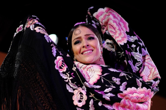 Internationally renowned flamenco dancer, Macarena Ramirez, dancing with a Spanish shawl (manton). She is holding the shawl up around her head with raised arms so that the shawl, which is black with large embroidered pink and white flowers, frames her beautiful smiling face. The background is dark, with a single spotlight.