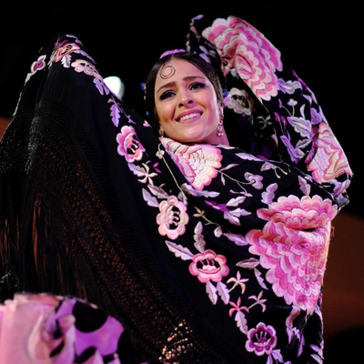 Internationally renowned flamenco dancer, Macarena Ramirez, dancing with a Spanish shawl (manton). She is holding the shawl up around her head with raised arms so that the shawl, which is black with large embroidered pink and white flowers, frames her beautiful smiling face. The background is dark, with a single spotlight.