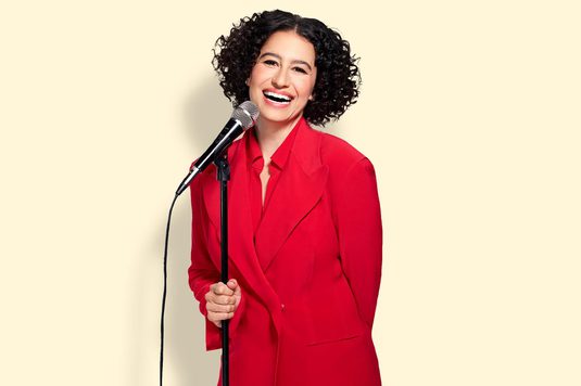 Ilana Glazer is wearing a red suit and stands confidently, holding a microphone, ready to speak or perform.