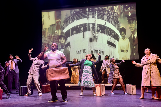 A group of people on stage dancing and signing while a picture of the Windrush ship is projected on the wall behind them